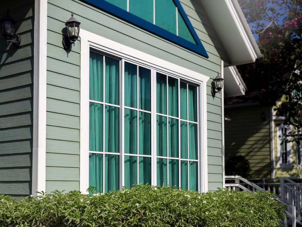 Sage green house exterior featuring a large, white-framed window with teal curtains and a lush bush in the foreground.
