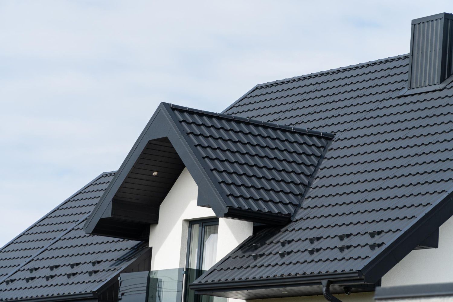 Black metal roof on a white house with a chimney and light blue sky in the background.