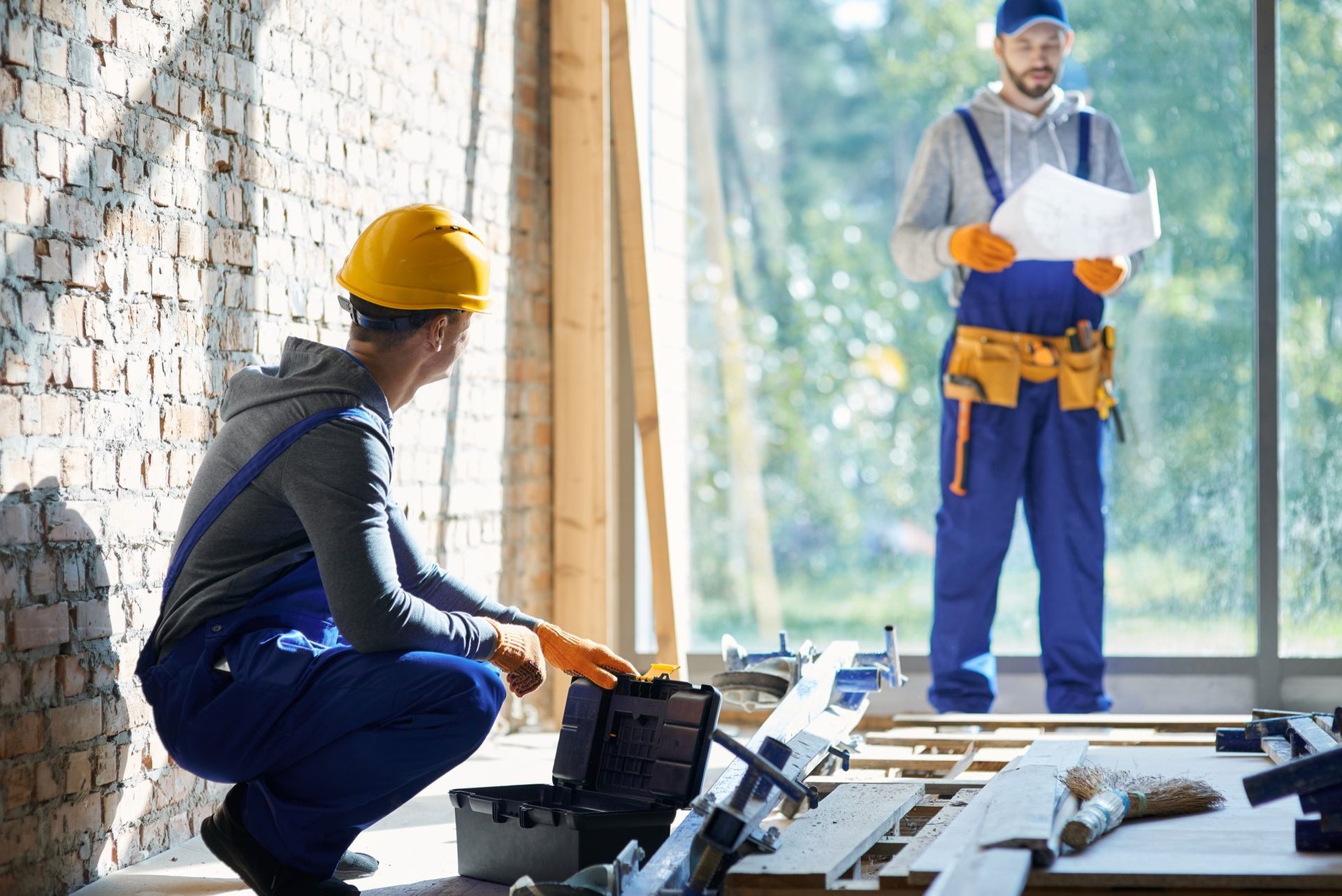 Two workers in blue overalls and hard hats at a construction site; one holds blueprints while the other opens a toolbox.