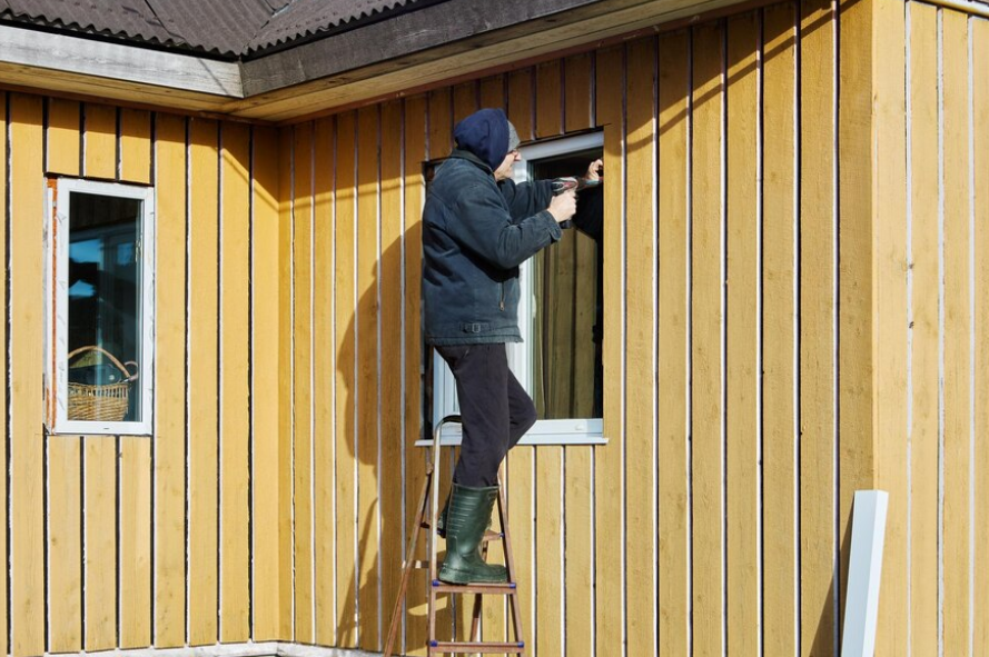 A person in a hooded jacket and boots stands on a ladder, using a tool to work on the trim of a window on a yellow house.