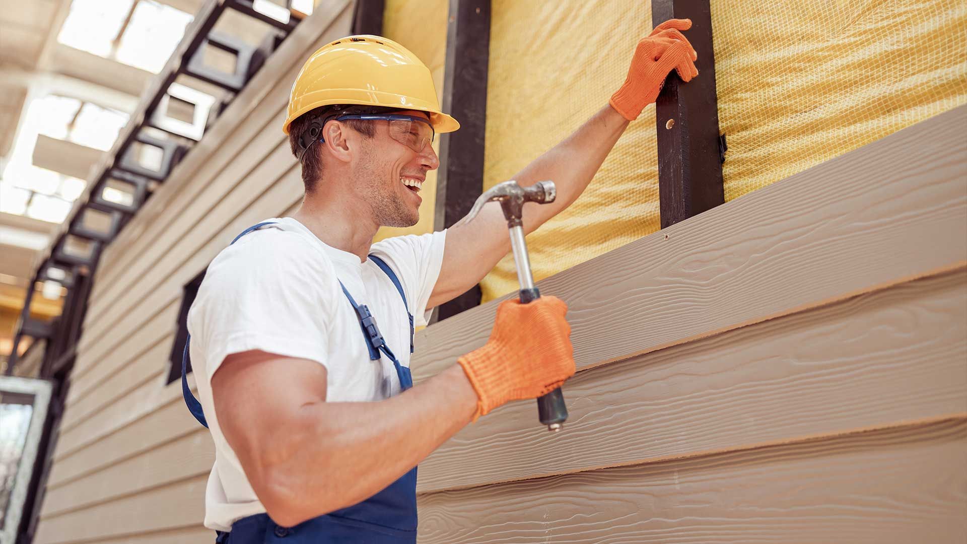 A construction worker in a yellow hard hat and orange gloves hammers siding onto an insulated building wall.
