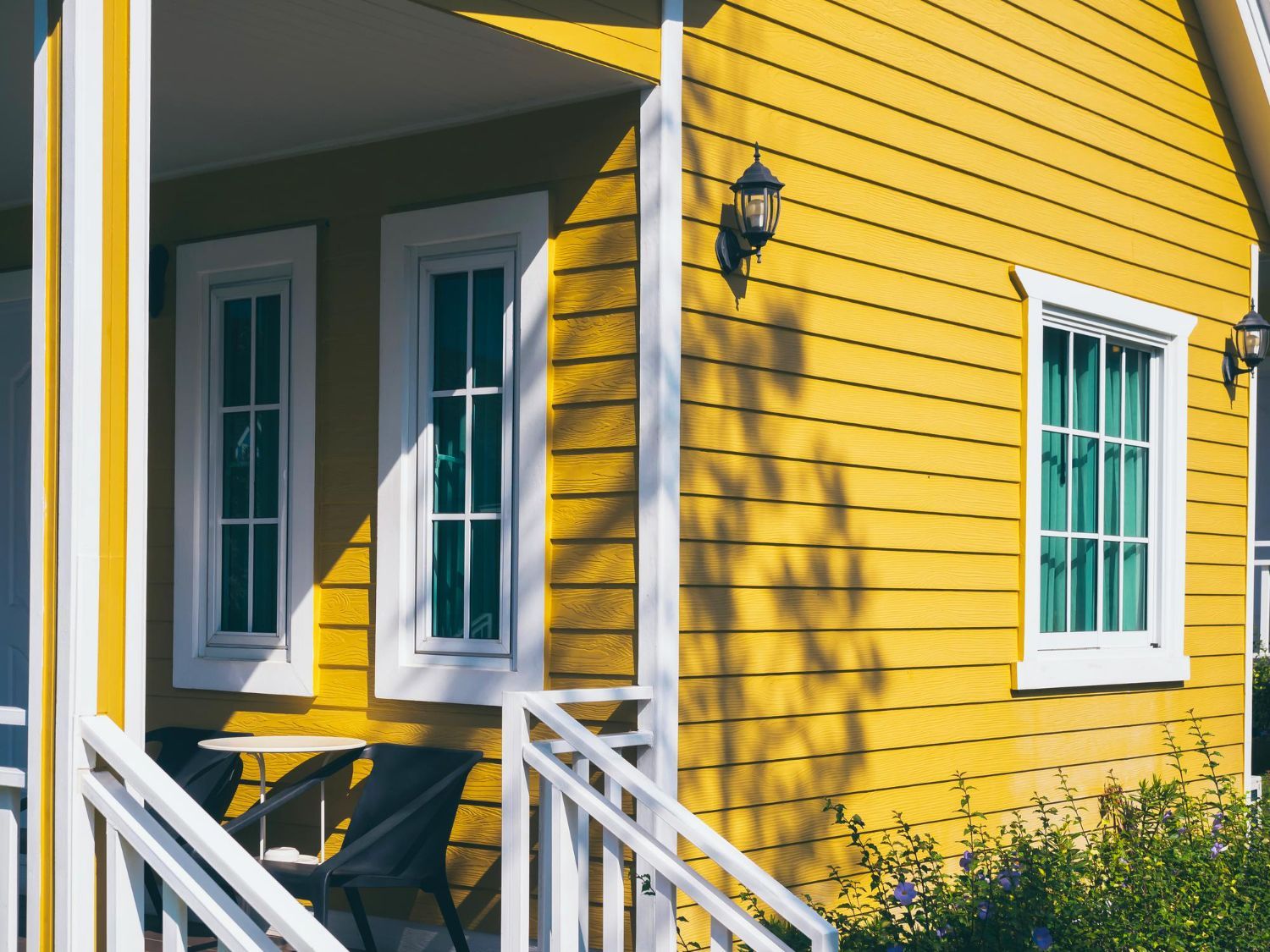 A bright yellow house with white trim, featuring a small porch with chairs and white railings under a sunny sky.
