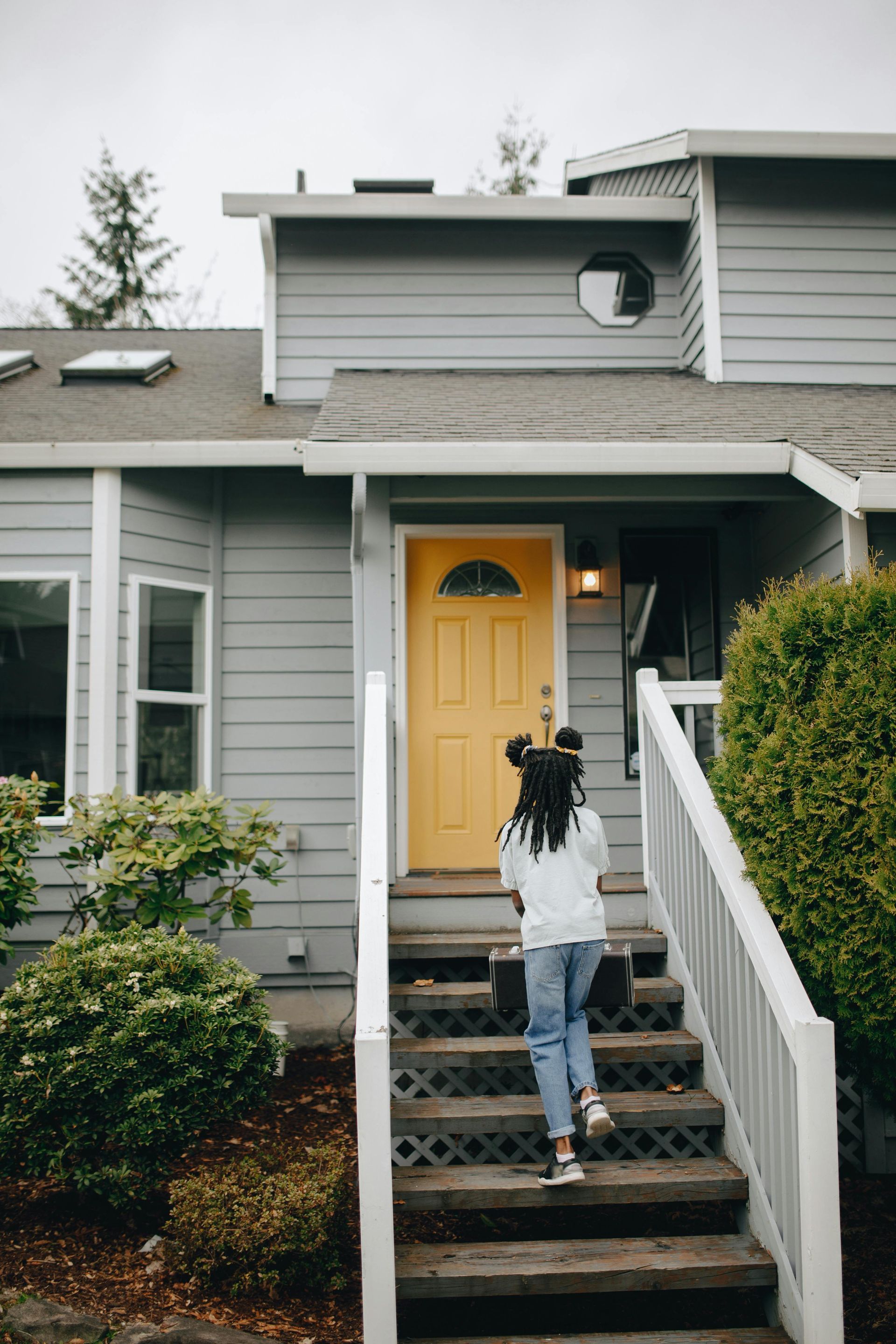 A person walking up stairs toward a yellow front door of a gray, multi-story house.